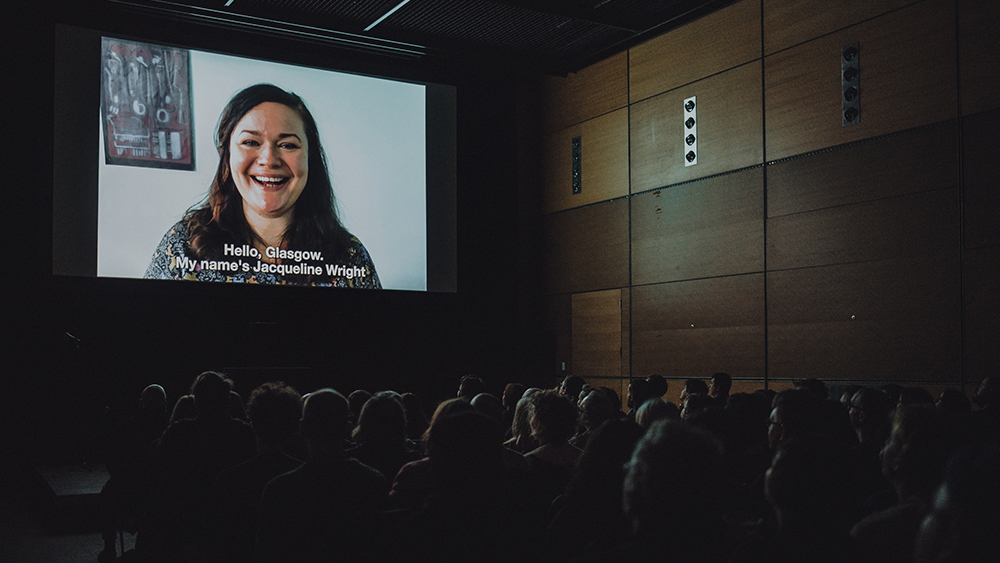 An audience sit in a cinema auditorium, facing a screen at the front on which a person is greeting them. The captions at the bottom of the screen read: 'Hello Glasgow. My name's Jacqueline Wright'.