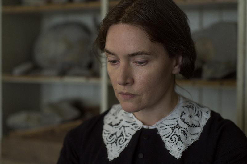 A person stands in a room, wearing a black dress with a white lace collar. Their eyes are cast slightly to the floor. In the background are some shelves with fossils gathered on them.