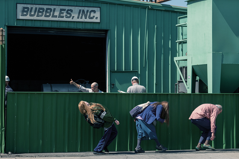 Three people walk crouched below a green metal wall, beyond which are three people on a worksite, two of which wear hard hats. A sign above the entrance reads Bubbles, Inc.