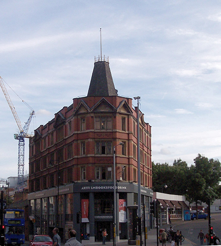 A red brick building which stands at the corner of a road in Manchester. A construction crane is in the background and a blue bus to the left, people wander the streets outside of the building.