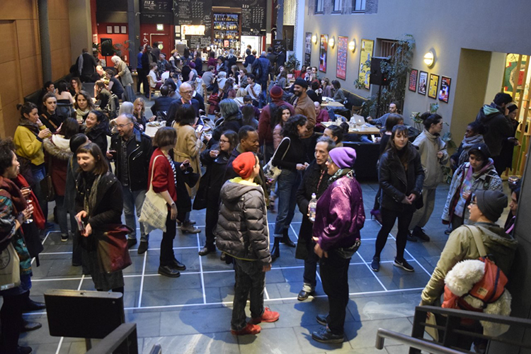 A crowd of people stand in a large cinema lobby, they stand in groups talking to each other.