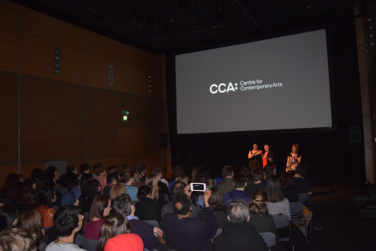 A crowd sit in a dark cinema auditorium. Three people stand at the front, underneath a screen showing a dark graphic with text which reads: CCA Centre for Contemporary Arts