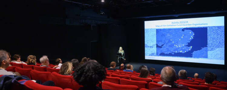 A large cinema auditorium. A woman on stage speaks to the audience. She has a map of the South East projected on the wall behind her.