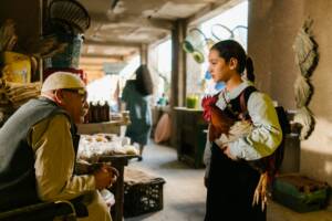 Still from THE PRESIDENT'S CAKE. A young girl talks to an old man in a market. She is holding a chicken under her arm.