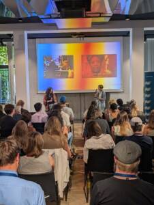 An audience listening to two speakers who are mid-conversation. Behind the speakers in a screen featuring brightly coloured projections.