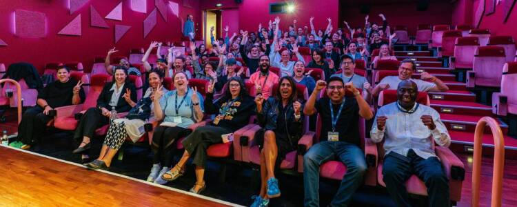 The attendees of the CICAE course in a large cinema. Their hands are raised and they smile at the camera. The lighting is a strong pink.