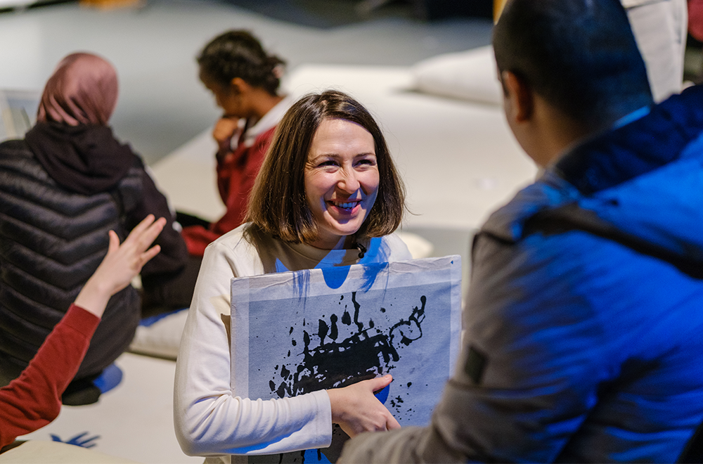 In workshop setting, a smiling person holds a black and white abstract splatter painting to their chest and smiles at a person in the foreground.