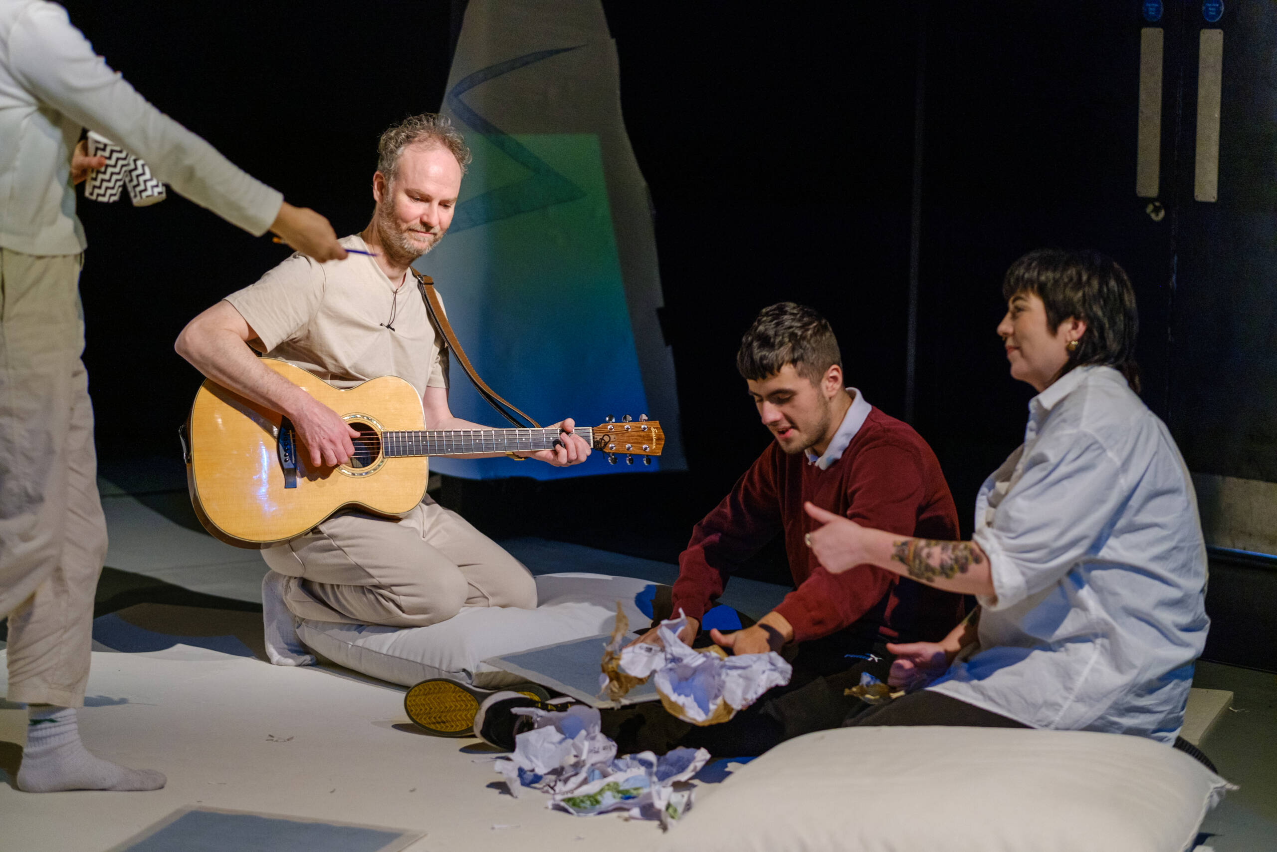 In a workshop setting, two people sit on the floor engaged in a craft activity. A person kneels near them on a cushion, playing a guitar.