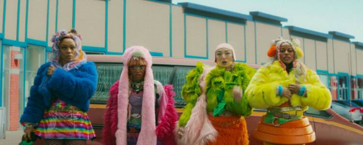 A still from I Love Boosters showing four women in brightly coloured clothing standing in front of a car in a carpark. Courtesy of Universal Pictures.