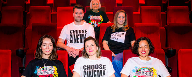 Six people seated in rows of bright red cinema seats, wearing ICO cinema-themed T-shirts. The shirts feature designs including 'Choose Cinema,' 'I'd Rather be in the Cinema' and 'It's Always Cinema Weather' printed in bold lettering. The photograph is taken inside an empty auditorium, with red seats filling the background.